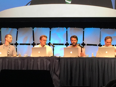 Four men sit at a table with laptops and microphones in front of them, facing an audience.