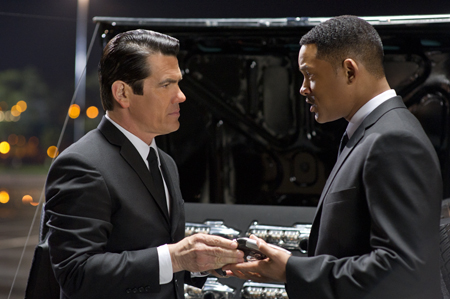 Two men in black suits stand facing each other near an open car trunk at night.