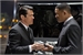 Two men in dark suits stand facing one another in profile against a dark background.
