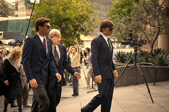 Two men in dark suits walk past a camera crew and trees in an outdoor urban setting.