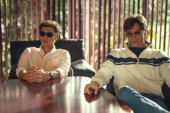 Two men in sunglasses sit at a dark wooden table in a room with vertical wooden slats in the background.