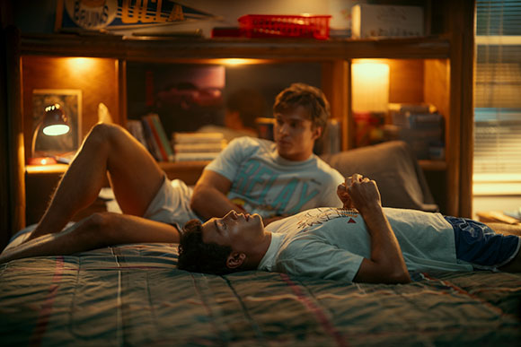 Two young men lie on a bed in a bedroom illuminated by warm lamp light.