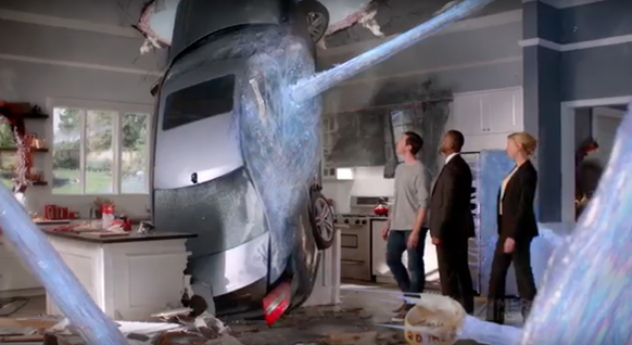 A gray car is stuck in a kitchen ceiling amidst debris and blue light beams, with three people watching.