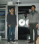 Two men stand in a lab next to a large black Scanity machine and server racks.
