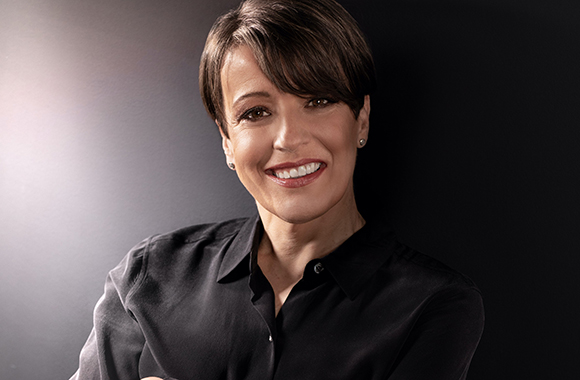 A smiling woman with short brown hair wearing a black button-down shirt against a dark background.