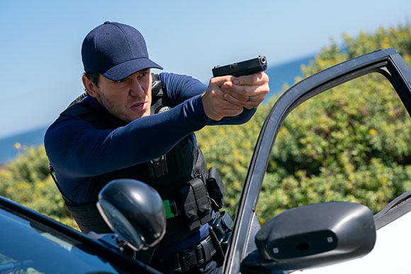 A man in a navy tactical vest and cap aims a handgun from a car door.