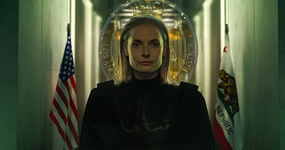A woman in black clothing stands between the United States and California flags in a dimly lit hallway.