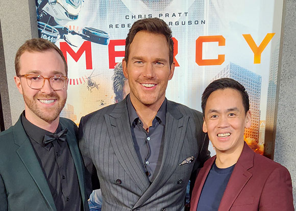 Three men smiling for a photo in front of a large movie poster for Mercy.