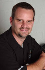 A man with short brown hair and a goatee smiling while wearing a black collared shirt.