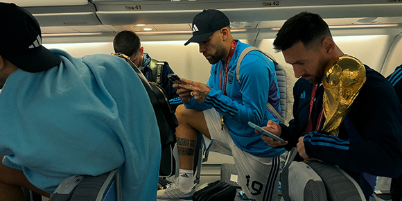 Soccer players in blue uniforms sitting on an airplane, one holding a gold trophy.