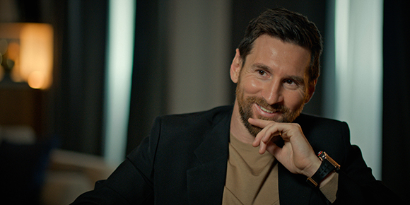 A smiling man with a beard wearing a black blazer and tan shirt, resting his chin on his hand.