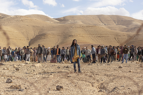 A man in a denim jacket stands in a rocky desert before a large crowd of people with mountains in the background.
