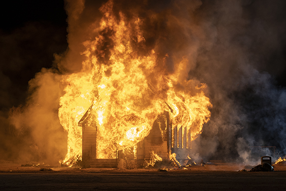 A wooden structure is completely engulfed in bright orange flames and thick clouds of smoke at night.
