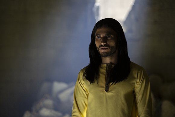 A man with long dark hair and a beard wearing a yellow tunic stands in a dark, smoky room.