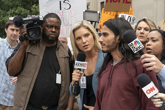 A man in a maroon hoodie is interviewed by several reporters holding microphones.