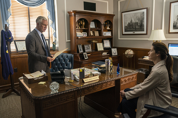 A man in a suit reads a paper to a seated woman in a wood-paneled office.