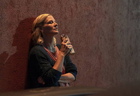 A blonde woman leans against a textured red wall, eyes closed, holding a bottle to her face.