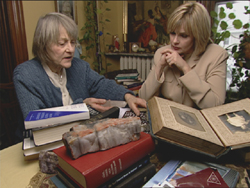 Two women sit at a table filled with books, looking at an open scrapbook.