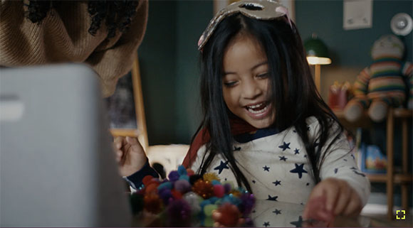 A young girl with long dark hair smiles while looking at a laptop and colorful pom-poms on a table.