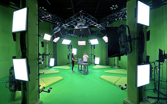 Two people stand in a green screen studio surrounded by bright studio lights and camera equipment.