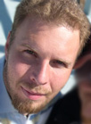 Close-up portrait of a man with light brown hair and a goatee looking toward the camera.
