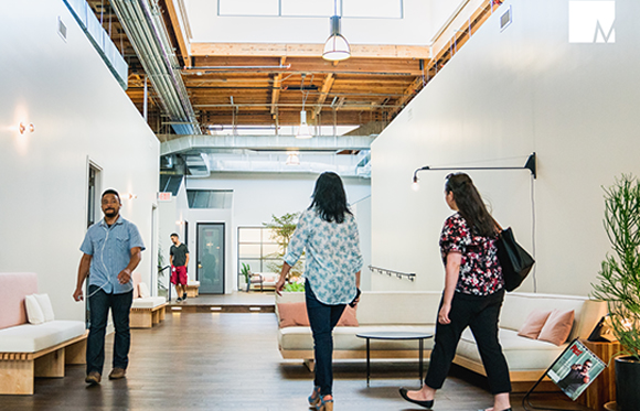 People walking through a bright, high-ceilinged modern office with white walls and wooden ceilings.