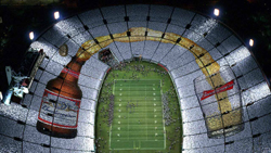 Thousands of people in stadium seating form a large image of a beer bottle pouring into a glass.