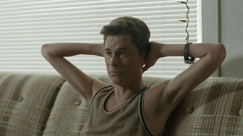 A man in a tank top sits on a patterned couch with his hands clasped behind his head.