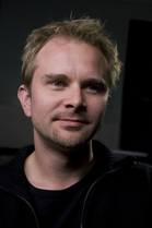A man with blond hair and light eyes looking slightly off-camera against a dark background.