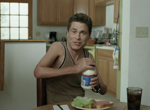 A man in a brown tank top sits at a table holding a bottle of mayonnaise in a kitchen.