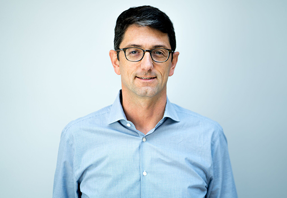 A man with dark hair and glasses wearing a light blue button-down shirt against a plain light background.