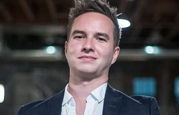 A man with short brown hair wearing a dark blazer over a white button-down shirt.