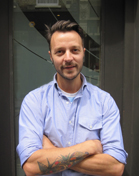 A man with dark hair and a light blue button-down shirt stands with arms crossed, showing a tattoo on his forearm.