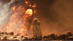 A person in a long cloak faces a colossal, fiery creature amidst smoke and debris.