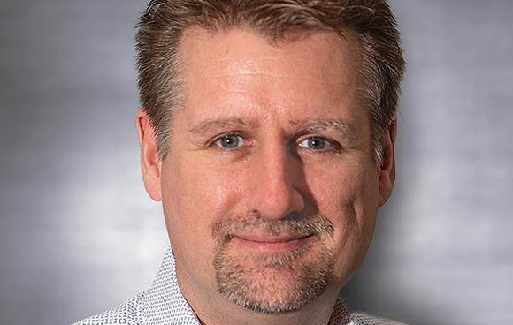 A man with light brown hair and a goatee smiling slightly against a gray background.