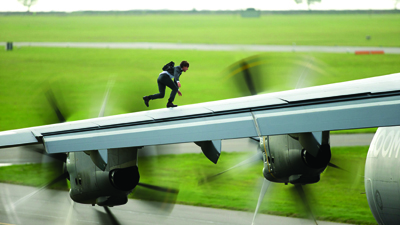 A person in dark clothing runs along the top of a large airplane wing with spinning propellers.