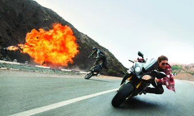 A motorcyclist leans into a turn on a paved road as a massive orange explosion erupts on a nearby hill.