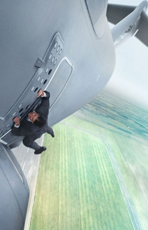 A man in a dark suit hangs from the belly of a large grey airplane flying over green fields.