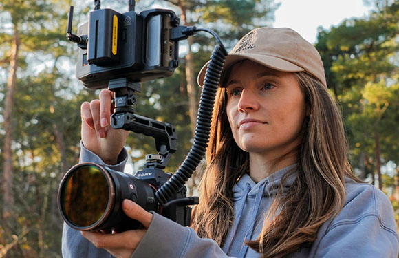 A woman in a tan cap and grey hoodie holds a professional camera mounted on a stabilizer rig outdoors.