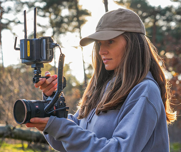 A woman in a baseball cap holds a professional camera rig with a monitor and wireless antennas in a forest.