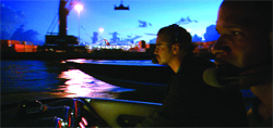 Two men sit on a boat at night under a dark blue sky with city lights in the distance.