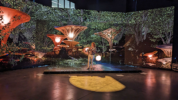 A person stands on a platform inside a room surrounded by large screens displaying a glowing forest scene.
