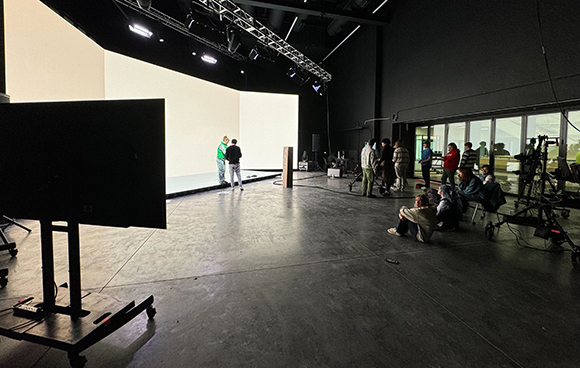 Film crew members standing and sitting in a large dark studio with a bright white backdrop.