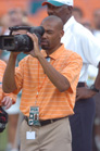 A man in an orange striped shirt holds a large professional video camera to his eye.