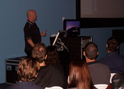 A bald man in a black shirt gestures while speaking to an audience seated in chairs. A screen displays an image behind him.