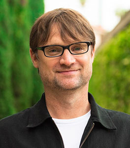 A man with short brown hair and black-rimmed glasses smiling against a blurred green background.