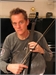 A man in a grey long-sleeved shirt holds a musical bow vertically in front of him.