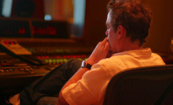 A man in a white shirt sits in a chair facing a large mixing console in a dimly lit studio.