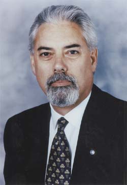 A man with grey hair, a grey goatee, wearing a black suit, white shirt, and patterned tie.
