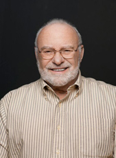 A smiling older man with glasses and a white beard wearing a striped button-down shirt.